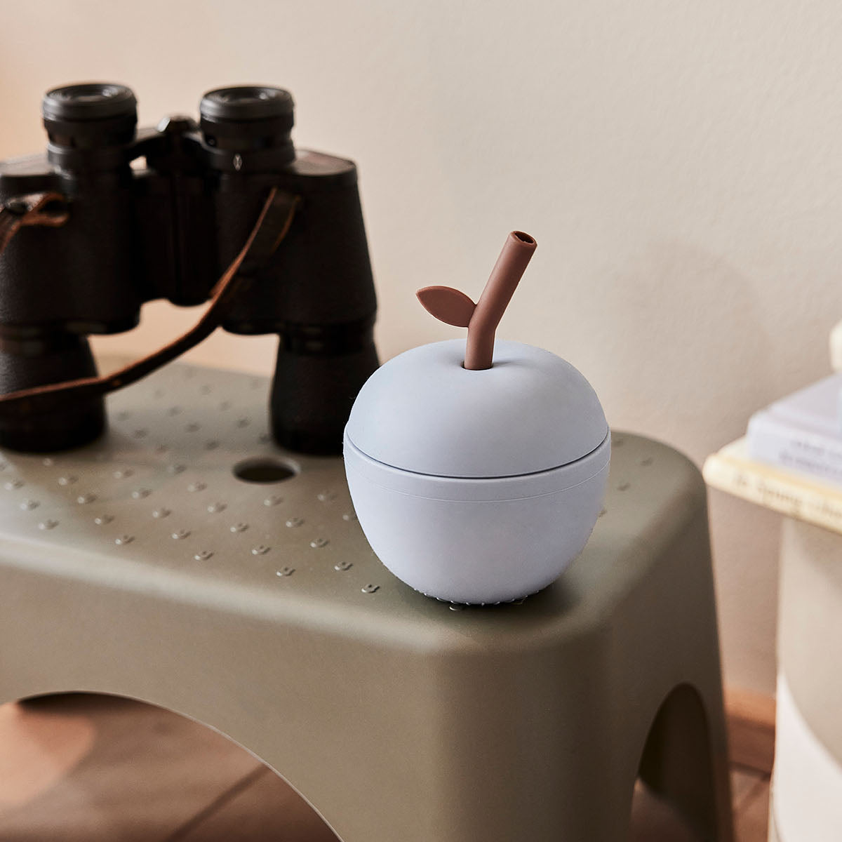 White ceramic apple-shaped container with a brown stem on a beige stool, with binoculars in the background.