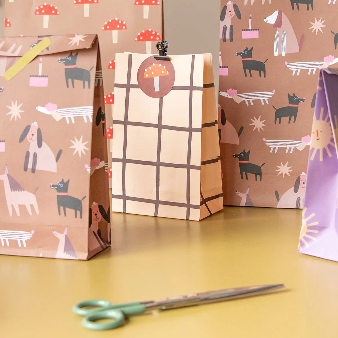 Several paper gift bags and wrapping paper sheets with whimsical dog and mushroom patterns, arranged on a yellow tabletop with a pair of green-handled scissors in front.
