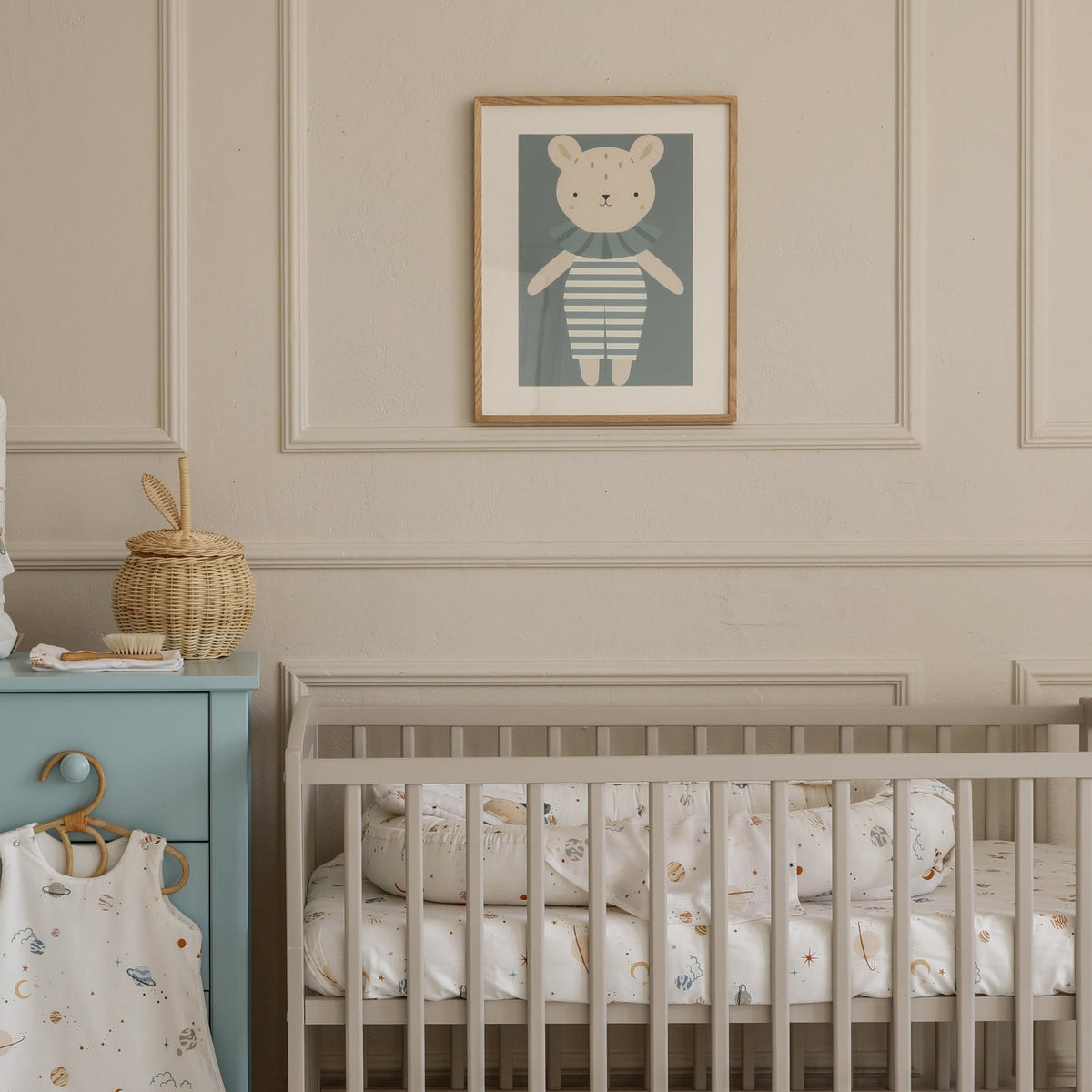 A styled nursery scene featuring a light beige wooden crib with a patterned mattress, a matching storage drawer beneath it, a light blue changing table with a patterned bag, and a framed bear illustration on the wall.