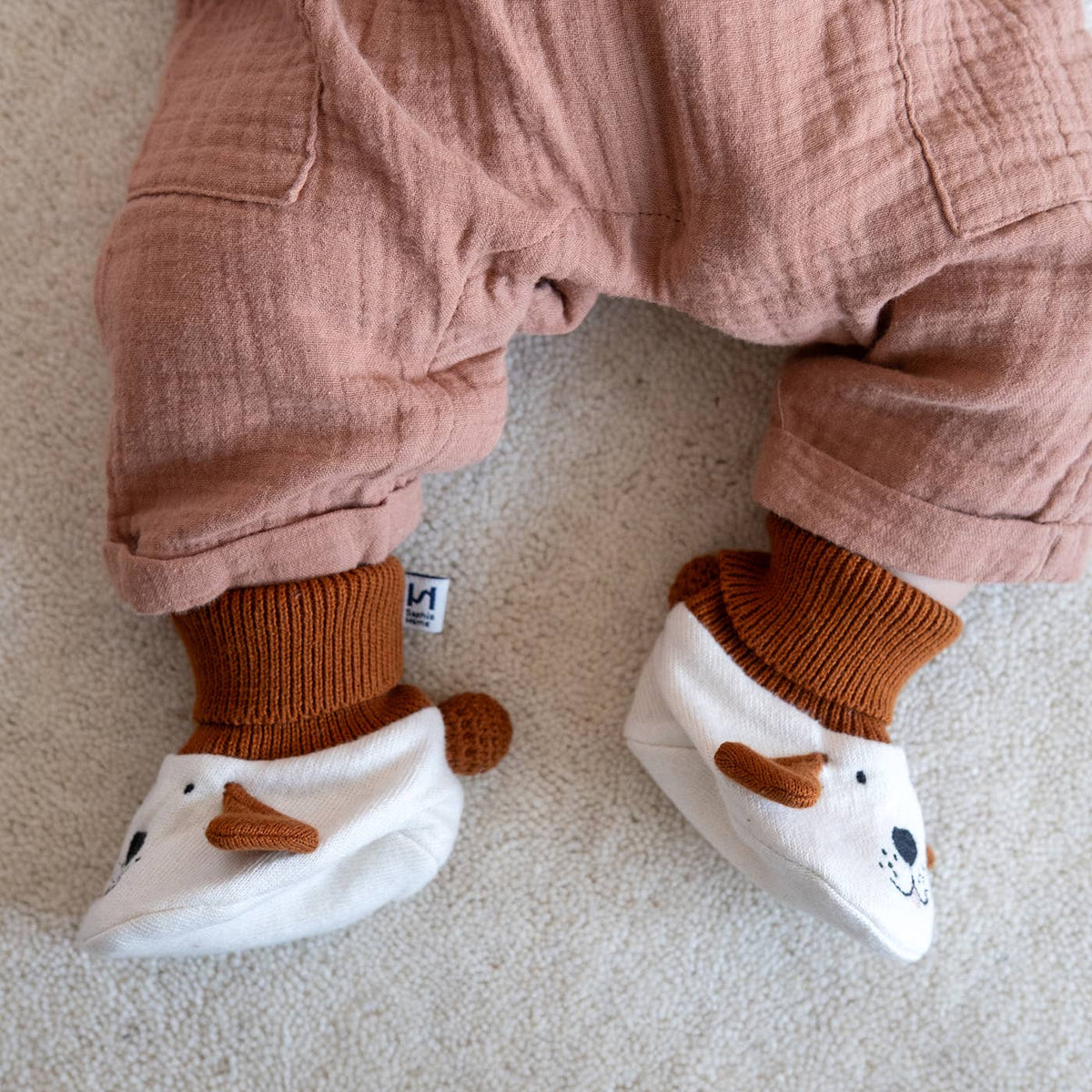 A close-up of a baby's legs wearing dusty rose muslin overalls, rust brown socks, and white bootie-style socks with brown ear details, laid on a light beige carpet.