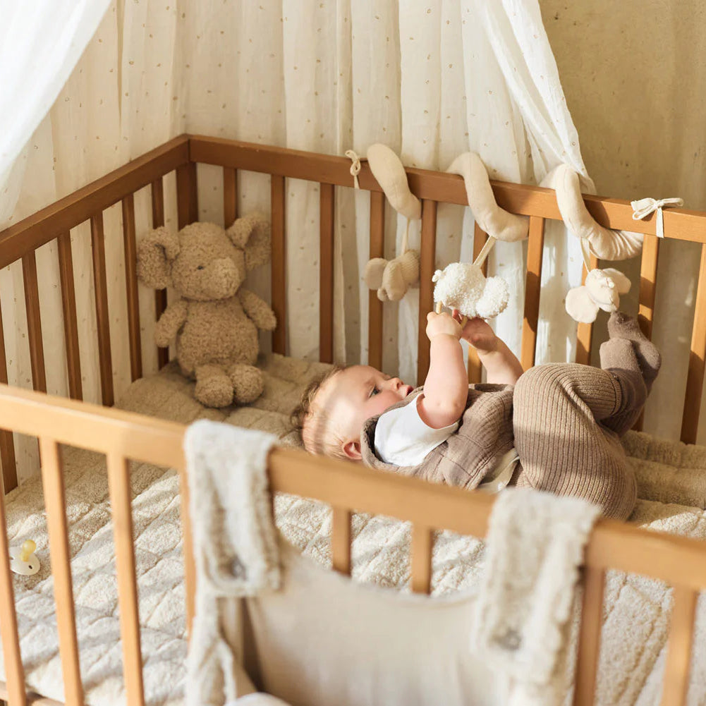 Baby lying in a wooden crib with toys and a teddy bear.