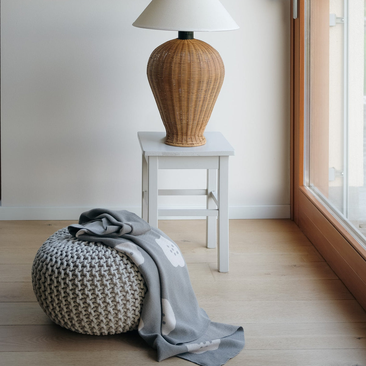 Woven lamp on a small table with a textured ottoman and blanket in a room with a window.