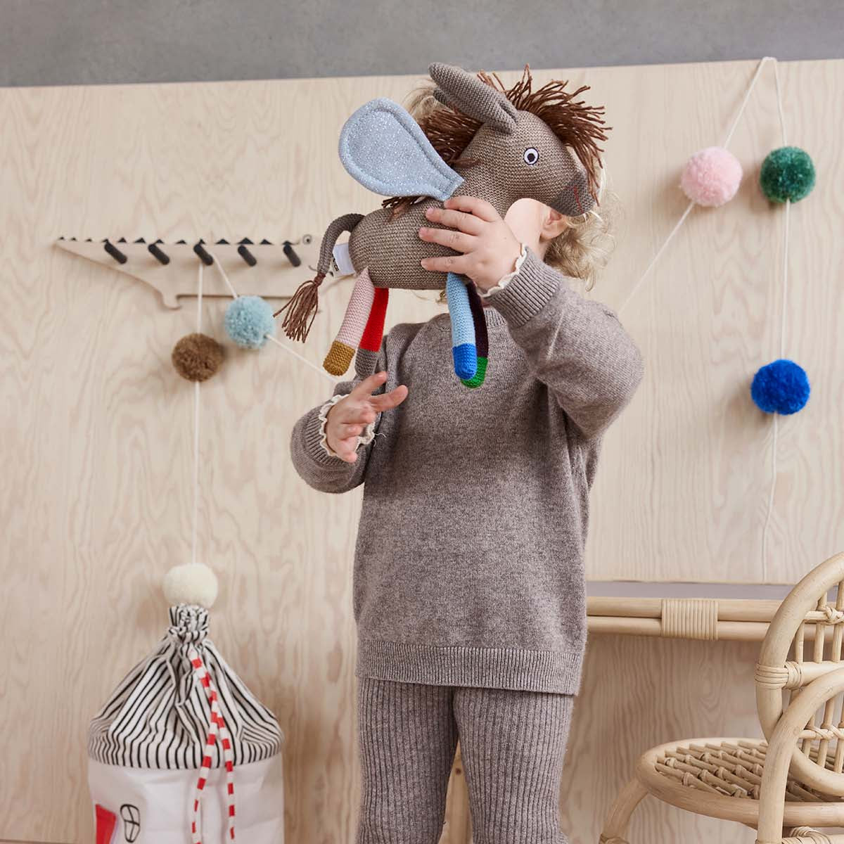 Child wearing a gray sweater and pants holding a toy horse in a room with a wooden floor and chair.