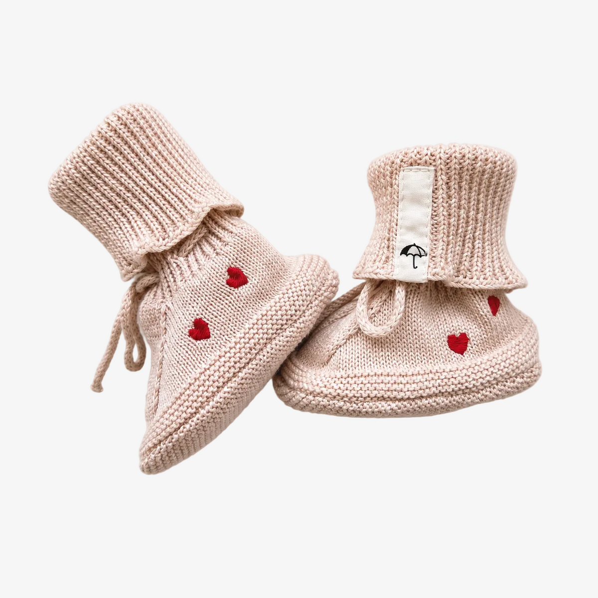 A pair of beige knit baby booties with small red heart details and a white brand tag featuring a black umbrella logo, displayed on a light cream background.